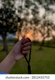 A hand holding a string of prayer beads, forming a silhouette with the tasbih against a warm, dramatic sunset in a tranquil garden, focusing on spiritual mindfulness and meditation.