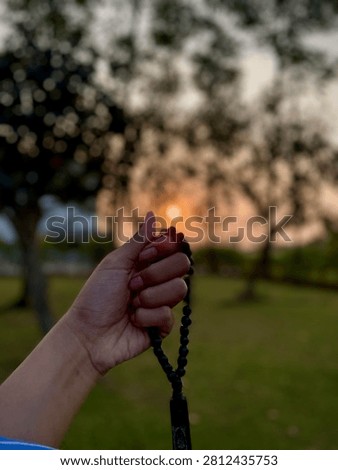 A hand holding a string of prayer beads, forming a silhouette with the tasbih against a warm, dramatic sunset in a tranquil garden, focusing on spiritual mindfulness and meditation.