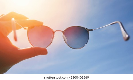 Hand holding stylish round sunglasses against bright sky and sun background. Wearing sunglasses on a sunny summer day. A man looks at the bright sun through polarized sunglasses. Summer mood - Powered by Shutterstock