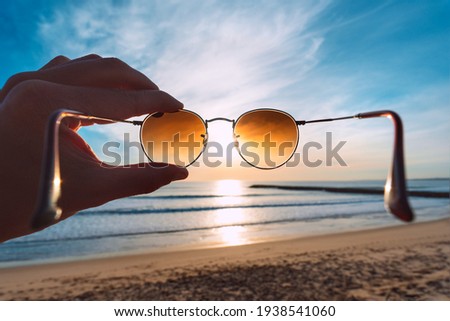 Hand holding stylish round sunglasses with brown lenses at sunset. Putting on sunglasses at sunny summer day near the ocean. Man looking at bright sun through polarized sunglasses. Summer vibes