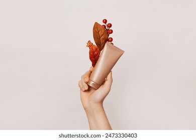 A hand holding a tan-colored cosmetic tube adorned with autumn-themed decorations including red berries and brown leaves - Powered by Shutterstock