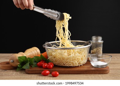 Hand holding tongs and cooked spaghetti from metal colander on wooden cuttingboard