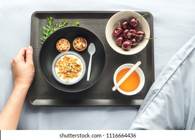 Hand Holding Tray Of Breakfast Set In Bed. Healthy Meal Such As Yogurt Muesli, Bean Snack, Honey, And Cherry In A Bowl Decoration With Leaves. Top Down View