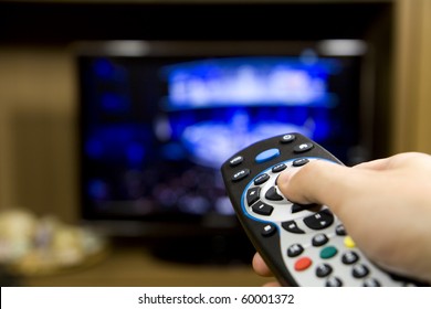 Hand Holding TV Remote Control With A Television In The Background. Close Up.