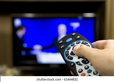 Hand Holding TV Remote Control With A Television In The Background. Close Up.