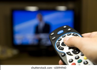 Hand Holding TV Remote Control With A Television In The Background. Close Up.