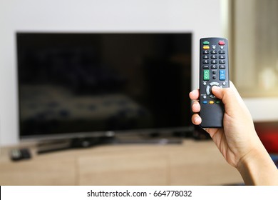 Hand holding TV remote control with a television in the background. 