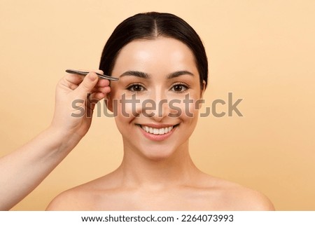 Hand Holding Tweezers And Shaping Eyebrows To Young Beautiful Indian Woman, Unrecognizable Beautician Plucking Brow Hairs Of Hindu Female Standing Over Beige Studio Background, Copy Space