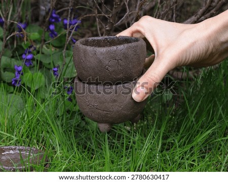 Hand holding two rustic brown handmade clay plant pots over lush green grass and spring flowers. Artisanal tripod pottery with raw cracked texture and organic earthy feel. Minimalist garden decor, small business pottery craft, and eco-friendly outdoor lifestyle concept.