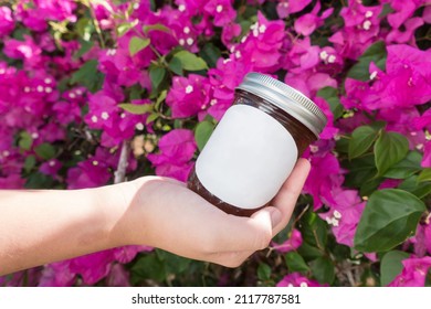 Hand Holding An Unlabeled Mason Jar Against A Floral Background. Jar Has A Custom Label Template Mockup To Be Replaced.