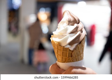 Hand Holding A Vanilla And Chocolate Soft Serve Ice Cream Cone Against Blurry Shop Of Ice Cream