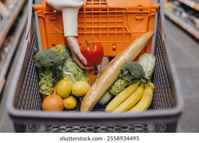 Hand holding a vibrant red bell pepper above a shopping cart brimming with fresh groceries in a bustling supermarket aisle - Powered by Shutterstock