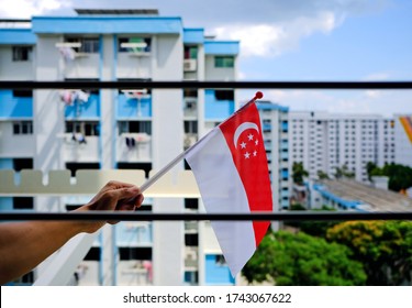 Hand Holding Waving Singapore Flag Outside HDB Flat Window. Due To Coronavirus Covid-19 Outbreak, The 2020 National Day Parade Would Be Broadcast Remotely, People'd Be Celebrating From Their Homes.