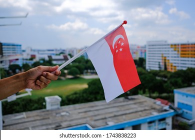 Hand Holding Waving Singapore Flag Outside HDB Flat Window. Due To Coronavirus Covid-19 Outbreak, The 2020 National Day Parade Would Be Broadcast Remotely, People'd Be Celebrating From Their Homes.