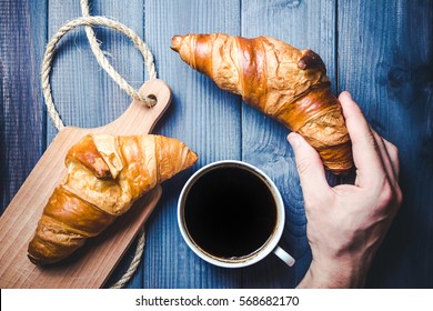 Hand Holding White Coffee Cup On Blue Wooden Table With Croissant.