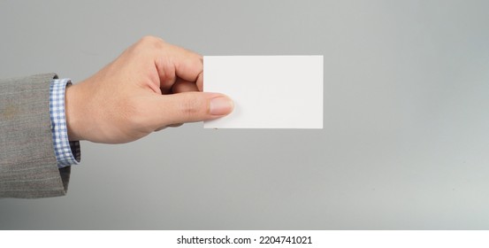 Hand Is Holding White Empty Card And Wear Suit On Gray Background. Business Man Concept. Copy Space For Text