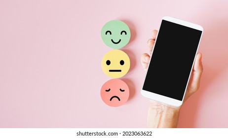 Hand Holding White Mobile Phone With Clipping Path On Touch Screen, Above Emotion Face On Sweet Pink Background For Satisfaction Survey ,feedback Rating , Mental Health Or Phycology Counselor