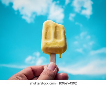 Hand Holding Yellow Frozen Popsicle Ice Pop Melting on Summer Blue Sky Background