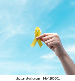 Hand Holding Yellow Gold Ribbon Awareness Over Blue Sky. Symbol For Support Suicide Prevention, Endometriosis, Sarcoma Bone Cancer, Bladder Cancer, Liver Cancer And Childhood Cancer Awareness Concept.