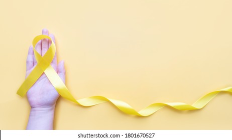 Hand Holding Yellow Gold Ribbon Awareness On Yellow Background. Symbol For Support Suicide Prevention, Endometriosis, Sarcoma Bone Cancer, Bladder Cancer, Liver Cancer And Childhood Cancer Concept.