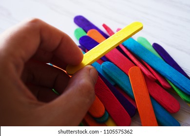 Hand Holding A Yellow Popsicle Stick Over  Its Pile During Process Of Selecting. The Stick Is Widely Used For Craft.