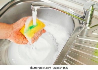 Hand holding a yellow scouring pad with a squirt of green soap on top making foam for washing dishes. Close-up of a man's hand performing housekeeping duties. Concept of cleanliness