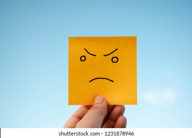 Hand holding yellow sticky note with unsatisfied face on blue sky background. Concept of unsatisfied or angry of someting