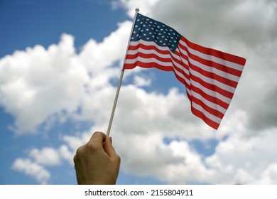 Hand Holds American Flag With Blue Sky And Clouds In Background.                           