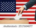 A hand holds a ballot in a ballot box against the background of the US flag. People cast ballots into a ballot box with the US flag in the background. Elections in the USA. Voting in the USA.