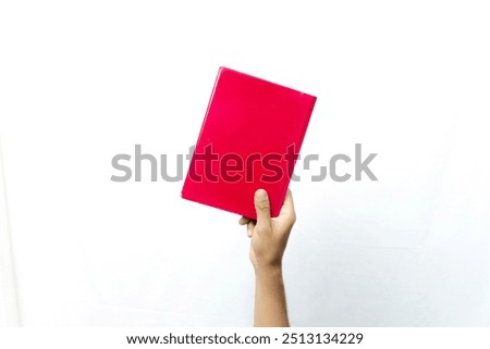 A hand holds a bright red book, against a plain white background. The book has a simple cover with a single uniform color.