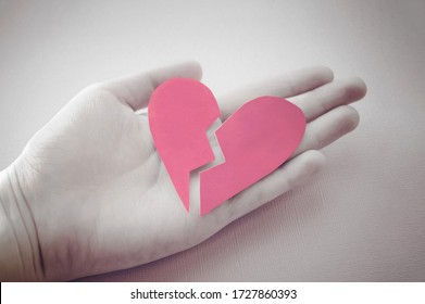 hand holds a broken heart. Grey and pink photo. Devorce and separation concept. Broken heart symbol. Paper heart. Relationship problems and lonliness. Two pieces of heart. Torn valentine card.