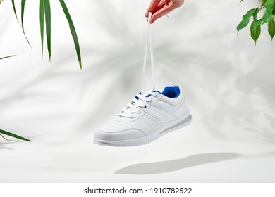 The hand holds by shoelaces white sneakers. Hand hold sporty shoes on a white background with palm leaves. Concept summer holiday.