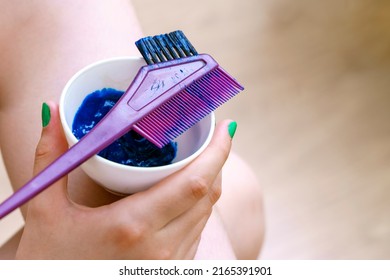 A Hand Holds A Cup Of Blue Hair Dye. On Top Is A Special Comb.