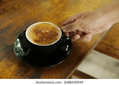 A hand holds a cup of creamy crème brûlée coffee on a wooden table. Perfect for use in coffee shop promotions, food blogs, and lifestyle content highlighting gourmet beverages. - Powered by Shutterstock