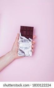 Hand Holds Dark Chocolate Bar In Foil On Pink Background