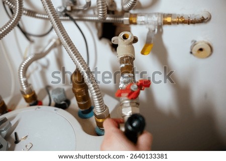 A hand holds a flashlight, illuminating section of water heater and its connected corrugated metal pipes, copper pipe, and valves against white wall, depicting focused plumbing repair or inspection.