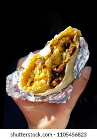 A Hand Holds A Foil-wrapped Carne Asada Burrito With A Bite Taken Out Of It Against A Dark Background. 