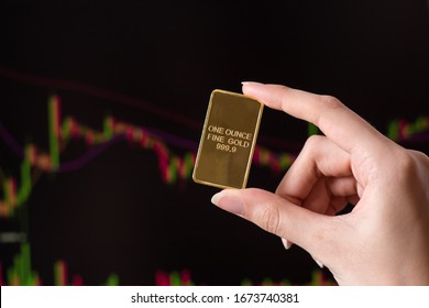 The hand holds a gold bar on the background of a laptop on which there is a trading price chart. One ounce of gold in hand. Trading gold on the stock exchange and Forex.