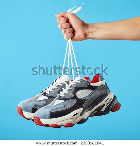 Hand holds hanging colored running sneakers by the laces on blue pastel background. Hand with a new casual shoe. Close up.