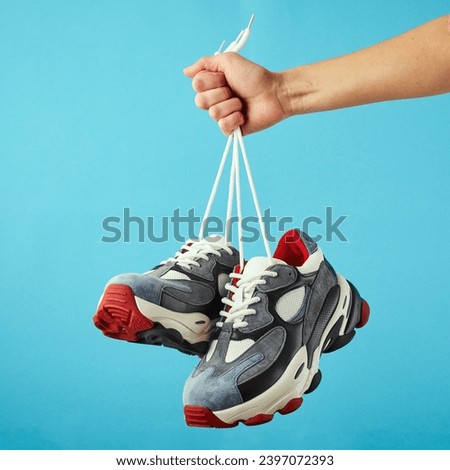 Hand holds hanging colored running sneakers by the laces on blue pastel background. Hand with a new casual shoe. Close up.