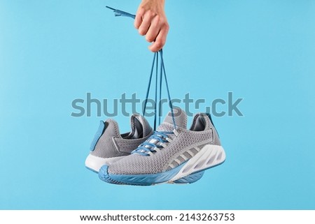 Hand holds hanging gray running sneakers by the laces on blue pastel background.  Hand with a new sport shoe. Stability and cushion running shoes. Close up.