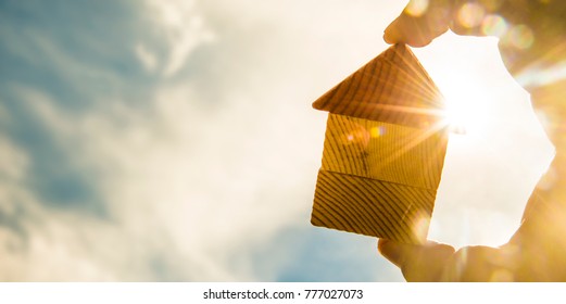 hand holds the last pieces of wooden house puzzle on blue sunny sky background. wooden home made from natural material of jigsaw. puzzle in the form of wood house. for sale or rent