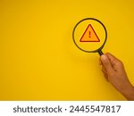 A hand holds a magnifying glass over a red warning sign with a yellow background. Perfect for illustrating attention, danger, alertness, caution, and warning concepts.