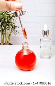 Hand Holds Medical Dropper With Liquid And A Drop Falling Over A Bottle Of Essence, Serum Or Care Oil In The Bathroom Against A Palm Tree. The Concept Of Face And Body Skin Care, Aromatherapy.