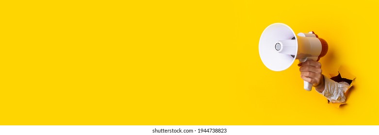 Hand holds a megaphone from a hole in the wall on a yellow background. Concept of hiring, advertising something. Banner. - Shutterstock ID 1944738823
