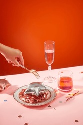 A Hand Holds A Metal Fork, A Star-shaped Balloon And Glittering Strings On A Plate, Next To A Glass Of Wine. Confetti On Pink Tabletop With Orange Background.