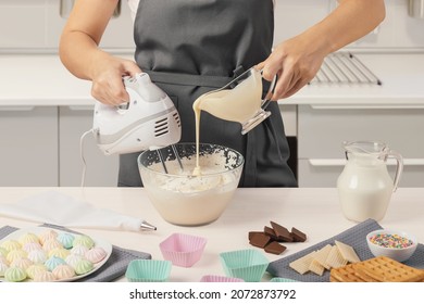 Hand holds a mixer and whips cream, the other pours milk. White kitchen. Ice cream, cream. Side view. On the table are towels, silicone molds, chocolate, a pastry syringe, sprinkles, waffles. - Powered by Shutterstock