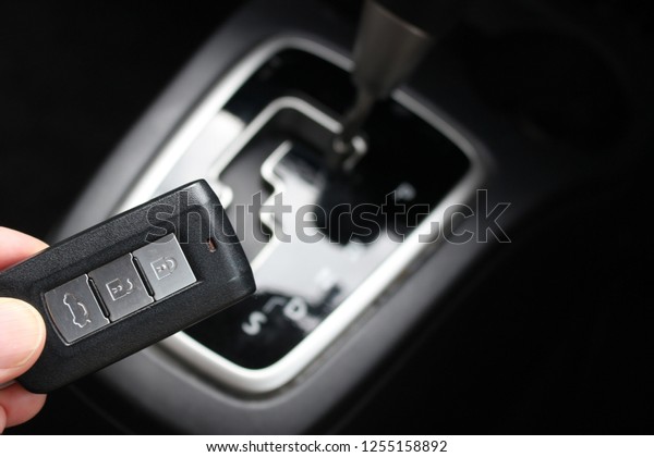 Hand holds a modern\
digital car key with an automatic transmission gear box visible in\
the background.