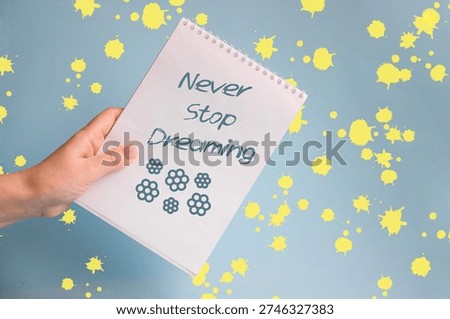 A hand holds a notepad with the text 'Never Stop Dreaming' against a blue background with yellow paint splatters.