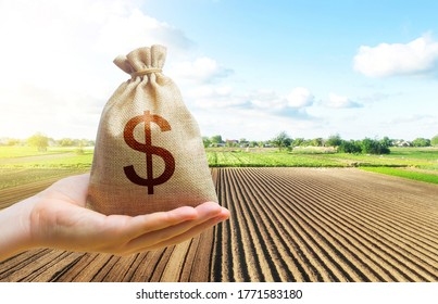 A Hand Holds Out A Dollar Money Bag On A Background Of A Farm Field. Lending Farmers And Agricultural Enterprises For Purchase Land And Seed Material, Equipment Modernization Support And Subsidies.